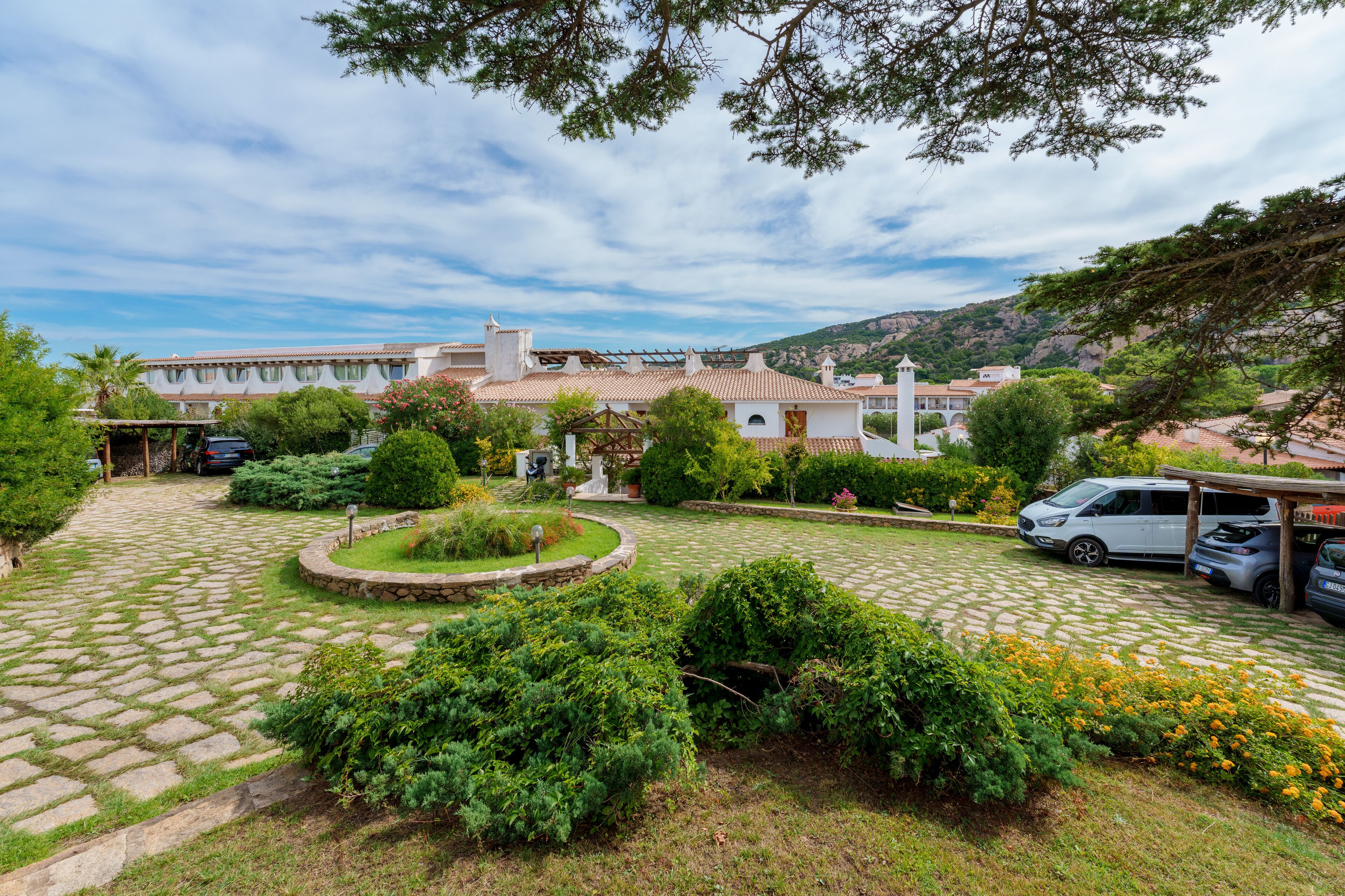 Une cour avec un chemin pavé, entourée de verdure et bordée d’un bâtiment doté d’un toit en tuiles.