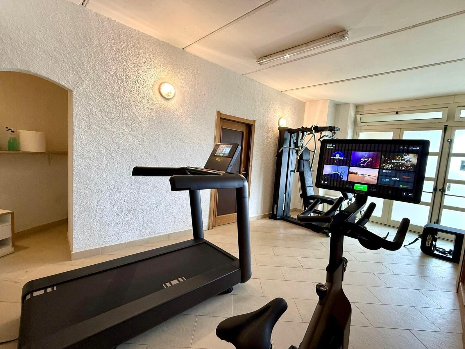 Un gimnasio en casa con cinta de correr, elíptica y bicicleta estática, una pantalla de televisión mostrando programas de entrenamiento y una puerta que da a otra habitación.