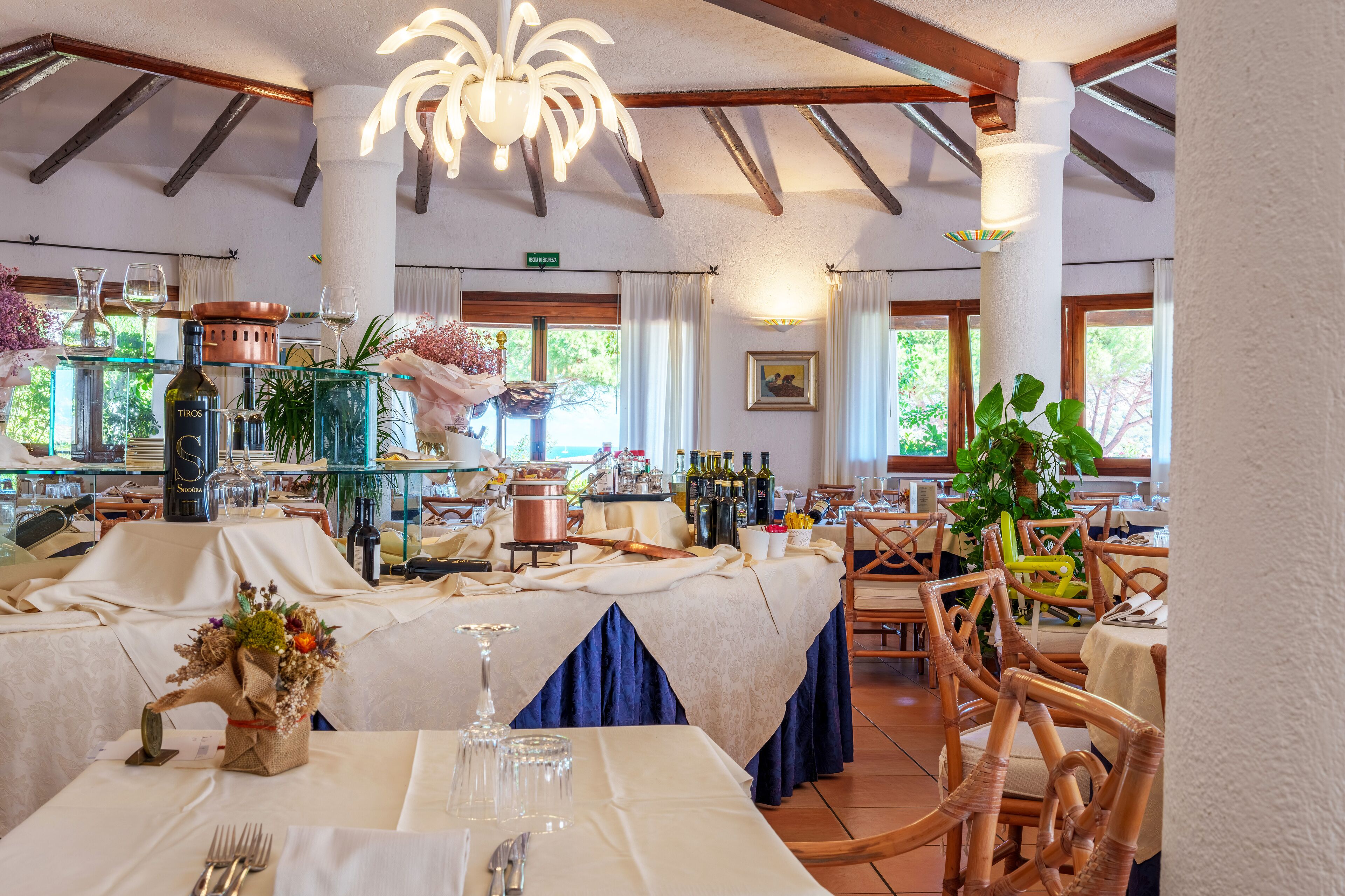 A dining area with tables set for a meal, a chandelier, and a view of the outdoors through large windows.