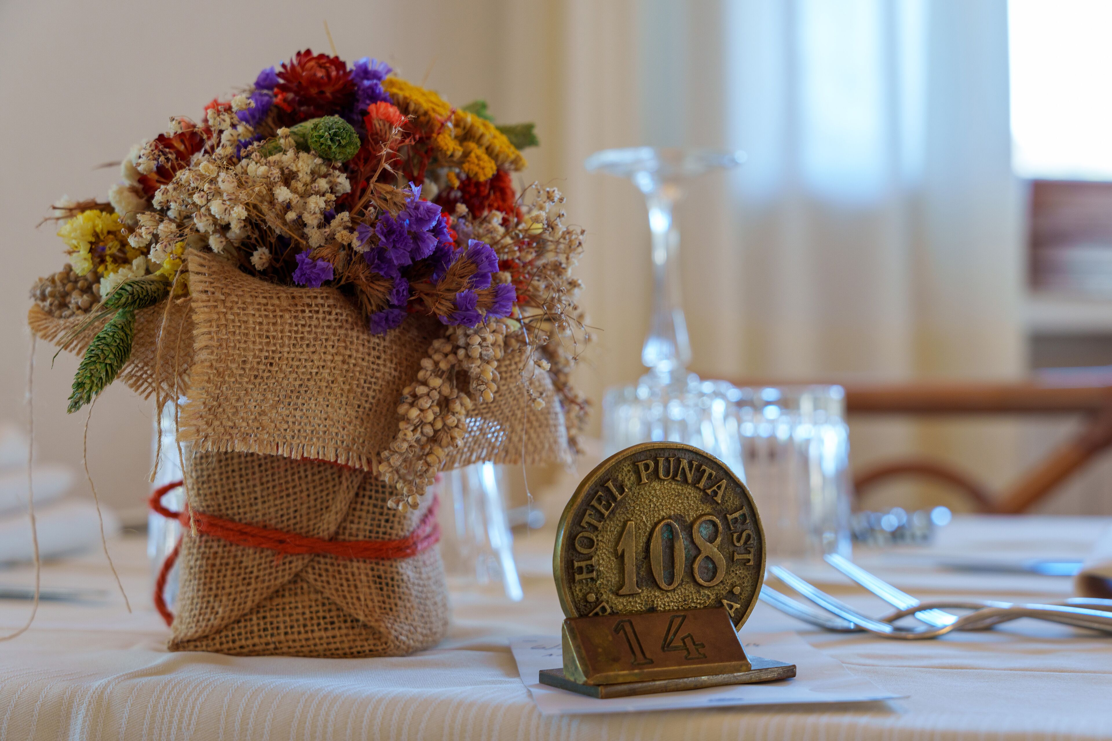Une table sur laquelle se trouve un sac en toile de jute rempli de fleurs colorées, une enseigne d’hôtel ancienne et un verre d’eau.