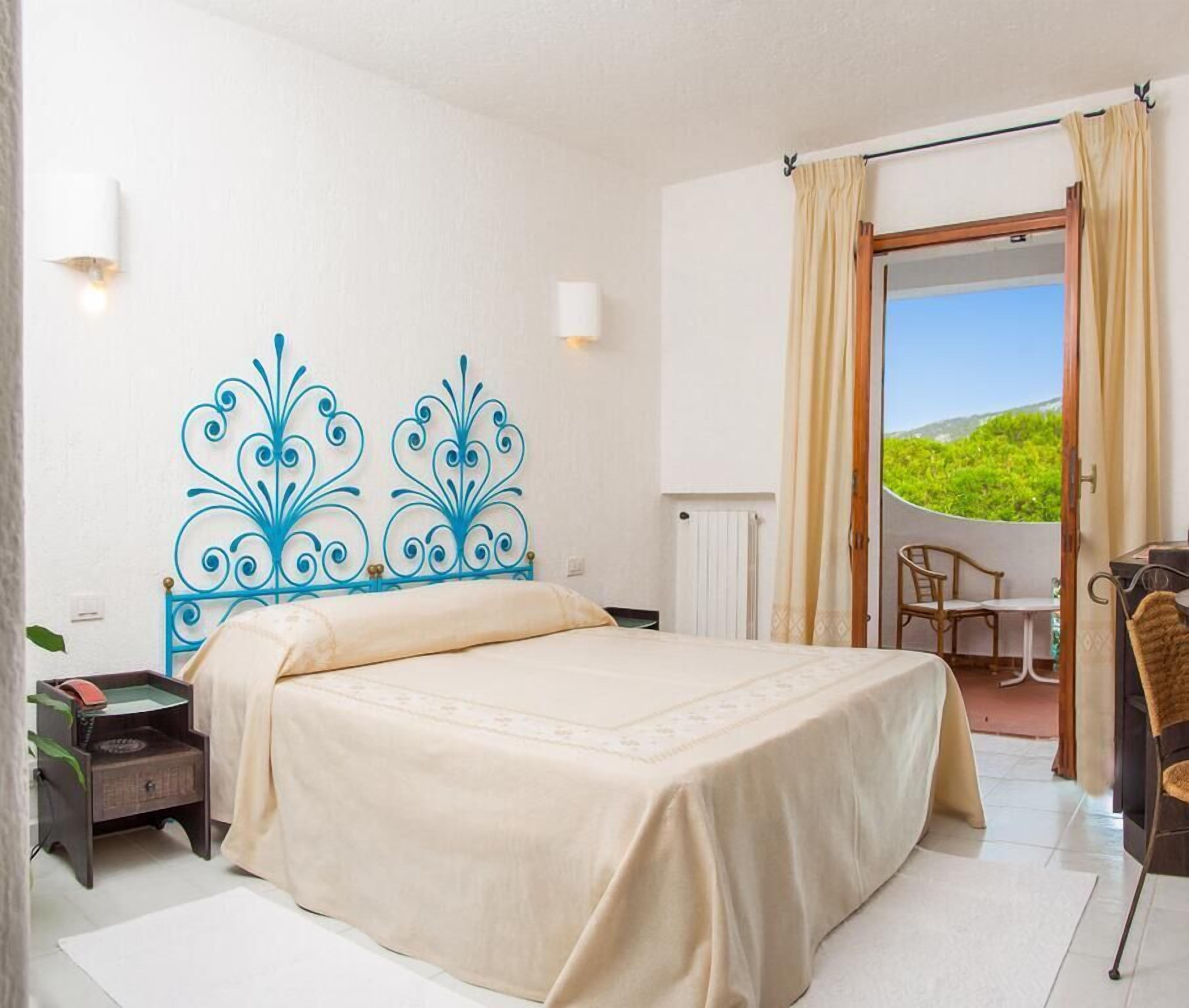 Ein Schlafzimmer mit Bett, Nachttisch, Sessel, Tisch und Blick auf Grünanlage durch eine Tür.