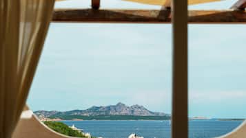 Une vue depuis une fenêtre, à travers un rideau, donnant sur un paysage côtier avec des montagnes et la mer.