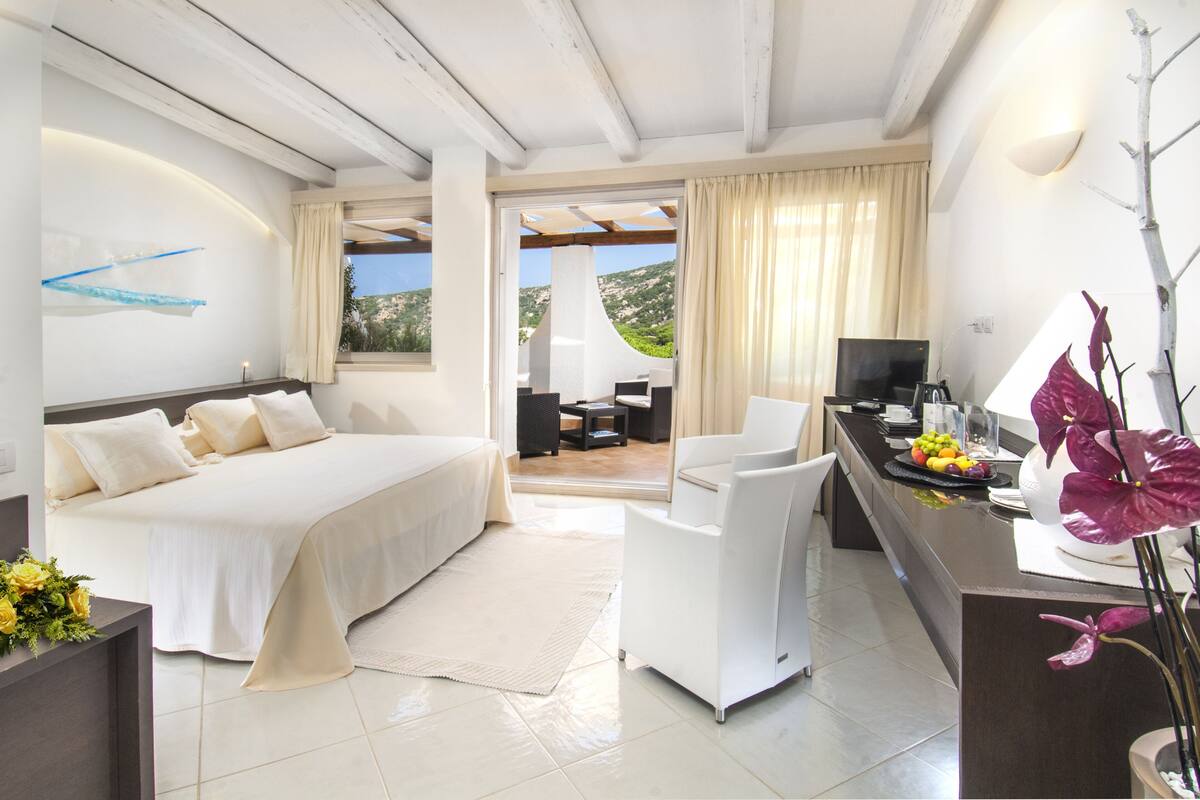 A modern hotel room with a bed, a desk, a chair, a TV, and a view of the outdoors.