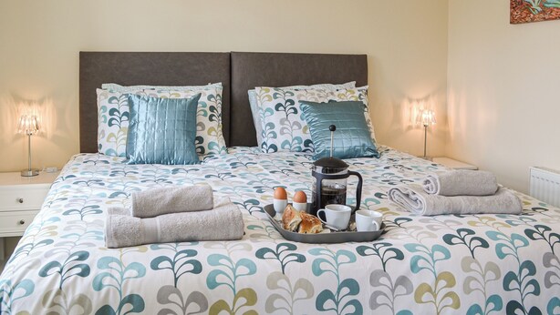 Un lit bien fait, recouvert d’une couvre-lit à motifs floraux, avec deux oreillers, un plateau contenant du pain, des œufs et une cafetière, et deux lampes de chevet.