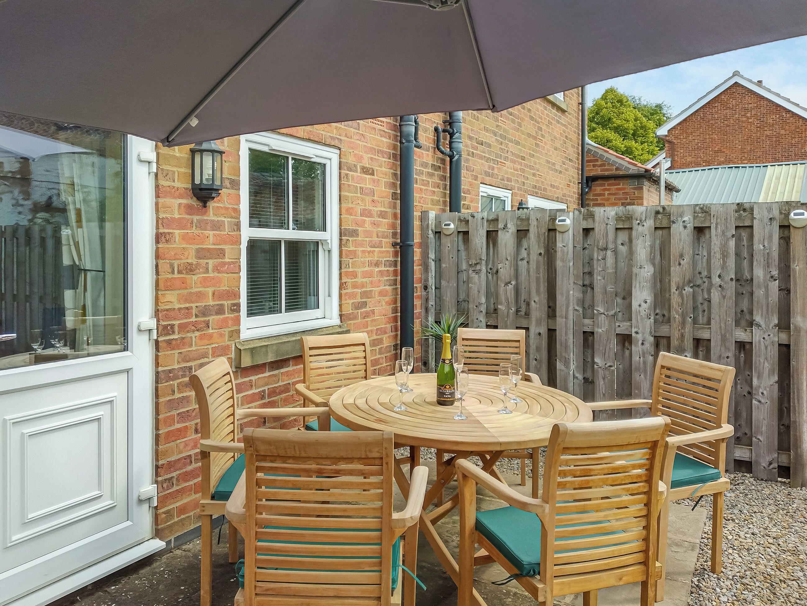 Eine Terrasse mit einem runden Tisch, sechs Holzstühlen, einer Champagnerflasche und einem Holzzaun im Hintergrund.