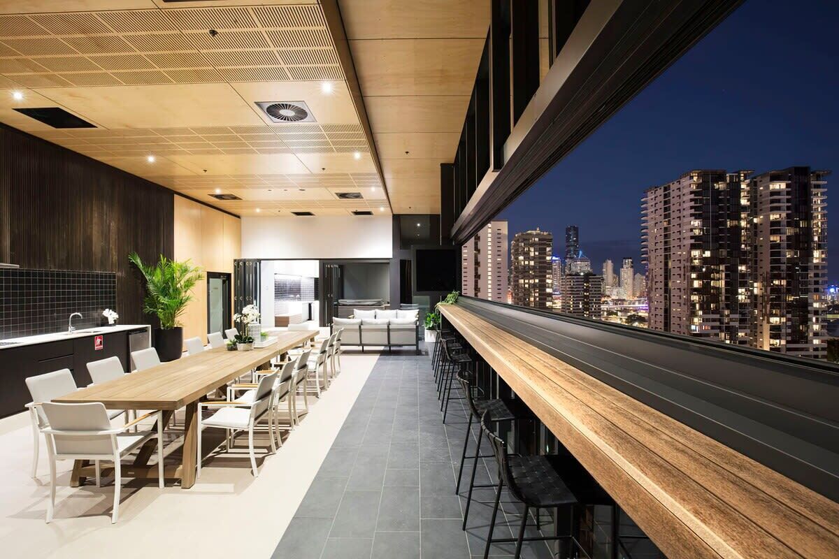 A modern dining area with a long wooden table and chairs, a kitchen area, and a view of a cityscape at night.