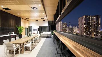 A modern dining area with a long wooden table and chairs, a kitchen area, and a view of a cityscape at night.