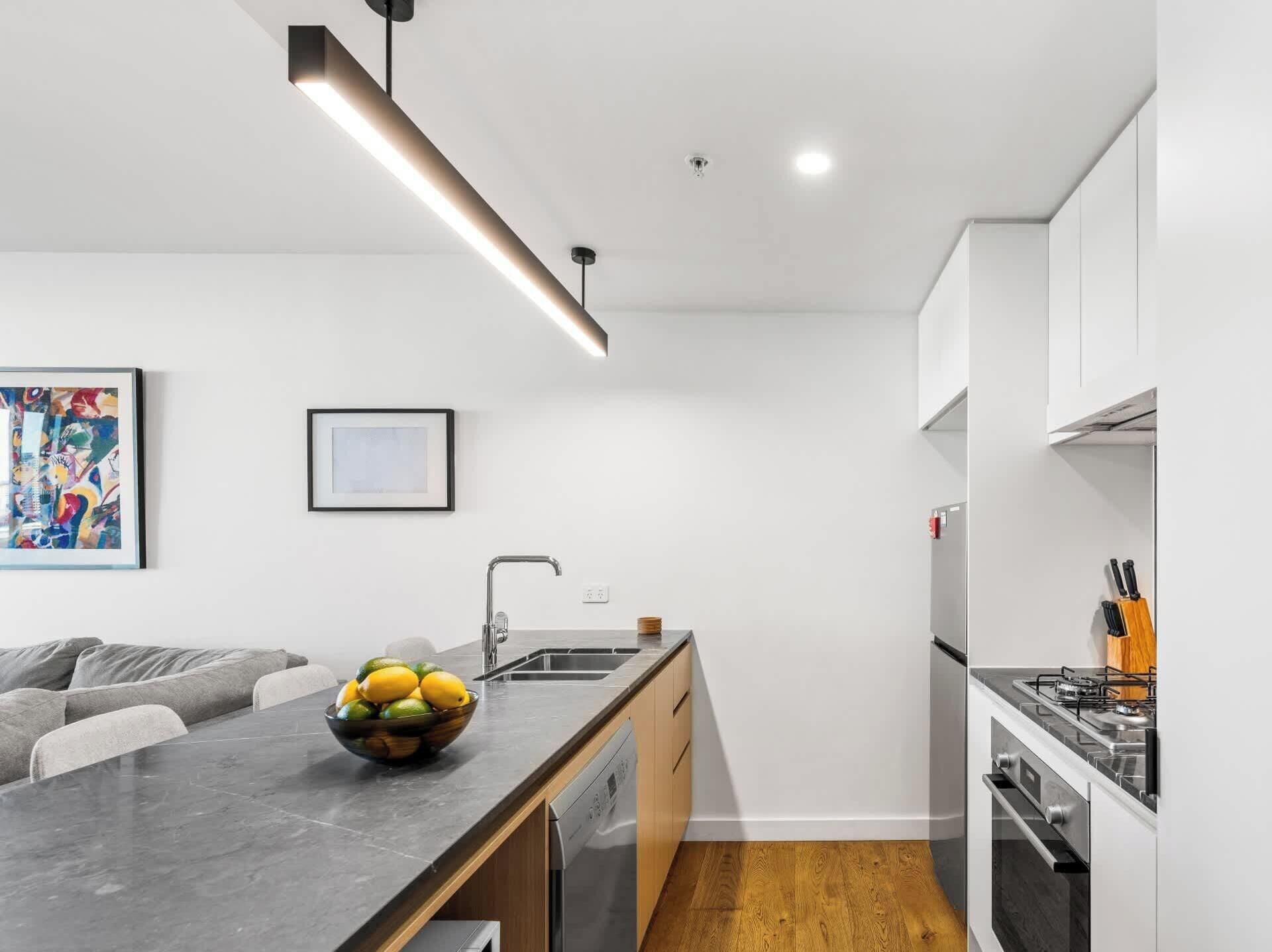 A modern kitchen with a built-in dishwasher, a gas stove, and a dining area with a marble table and a bowl of fruit.