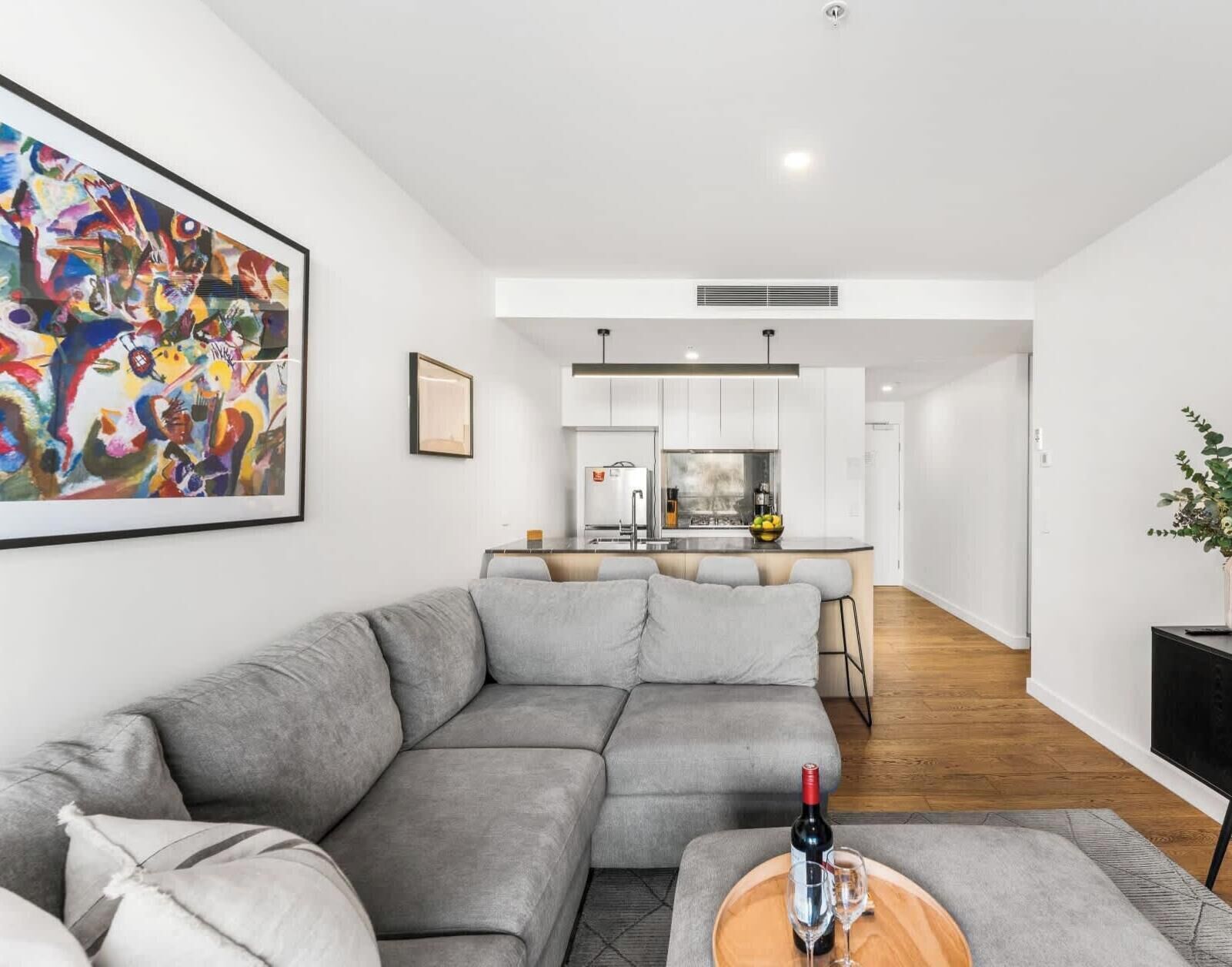A modern living room with a grey sectional sofa, a wooden coffee table with a wine bottle and glass, and a colorful abstract painting on the wall.