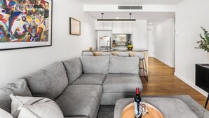 Ein modernes Wohnzimmer mit einem grauen Ecksofa, einem Couchtisch aus Holz mit einer Weinflasche und einem Glas sowie einem farbenfrohen abstrakten Gemälde an der Wand.