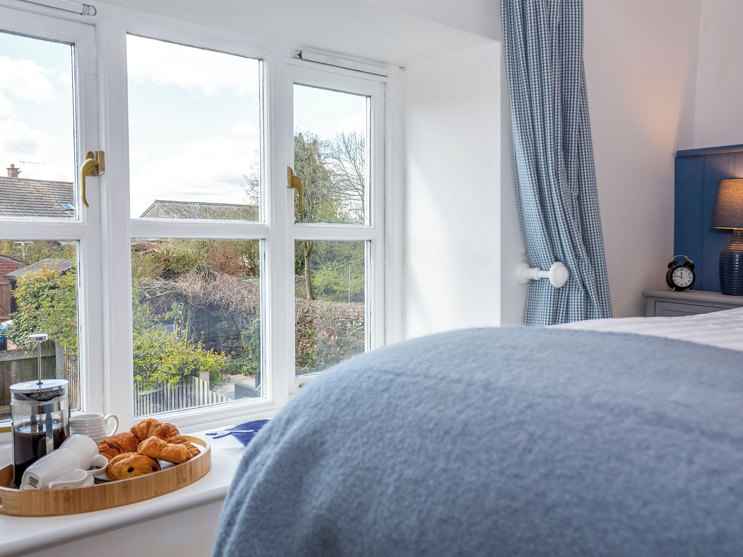 A bedroom with a bed, a bedside table with a lamp, a window with curtains, and a breakfast tray on the windowsill.