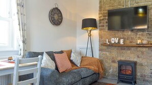 Ein gemütliches Wohnzimmer mit grauem Sofa, einem Holztischchen, einem Flachbildfernseher an einer Ziegelwand und einem Kaminofen.