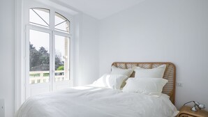 Ein Schlafzimmer mit großem Fenster, einem Bett mit weißer Bettwäsche und einem Weidenholz-Nachttisch mit Lampe.