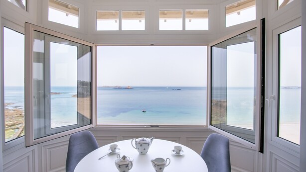 Ein Zimmer mit einem runden Tisch, gedeckt für ein Kaffeetrinken, großen Fenstern mit Meerblick und einer hellen Einrichtung.