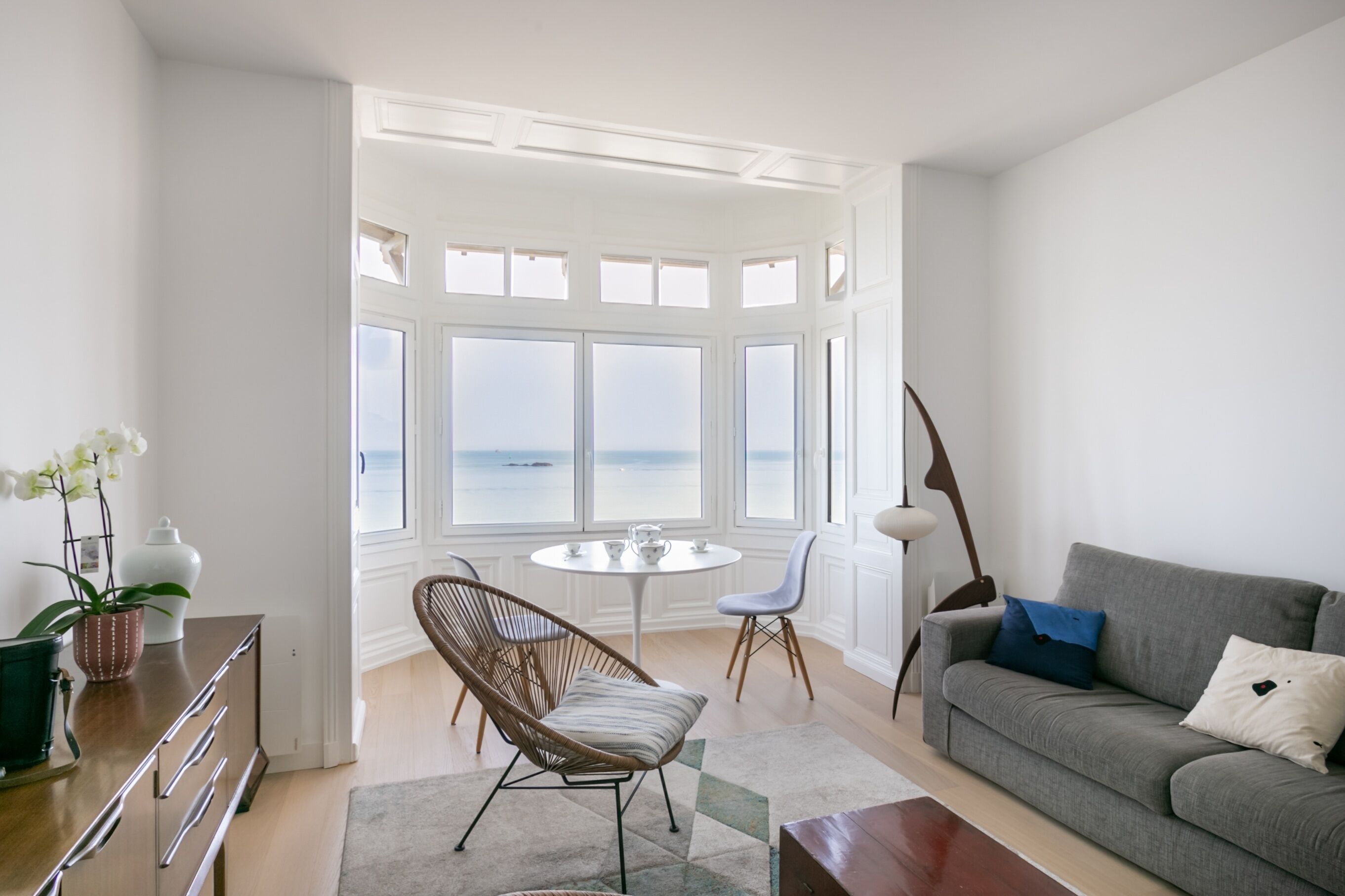 Ett modernt vardagsrum med utsikt över havet, en grå soffa, en fåtölj i rotting, ett runt bord och en lampa.