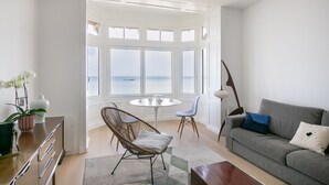 Ein modernes Wohnzimmer mit Meerblick, ein graues Sofa, ein Korbstuhl, ein runder Tisch und eine Lampe.