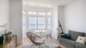Ett modernt vardagsrum med utsikt över havet, en grå soffa, en fåtölj i rotting, ett runt bord och en lampa.
