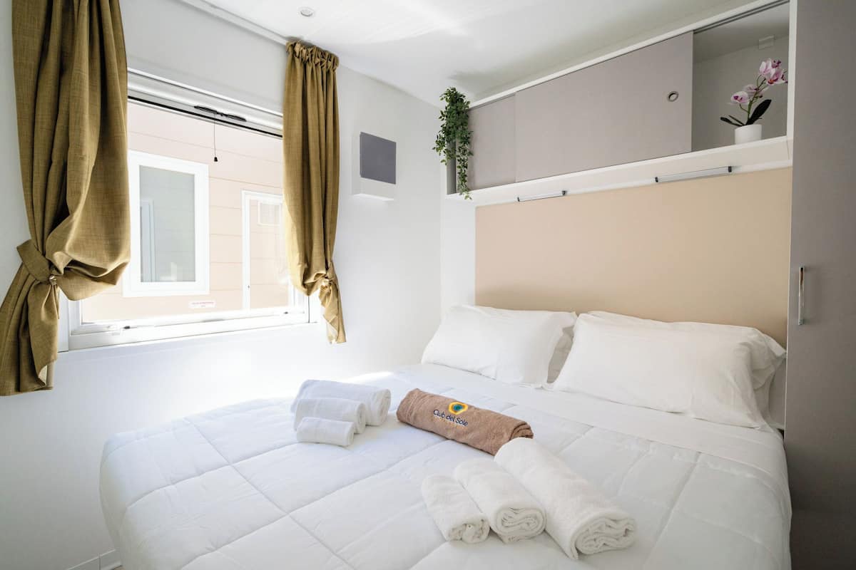 A hotel room with a bed, white bedding, a window with curtains, a bedside plant, and a towel with a logo.