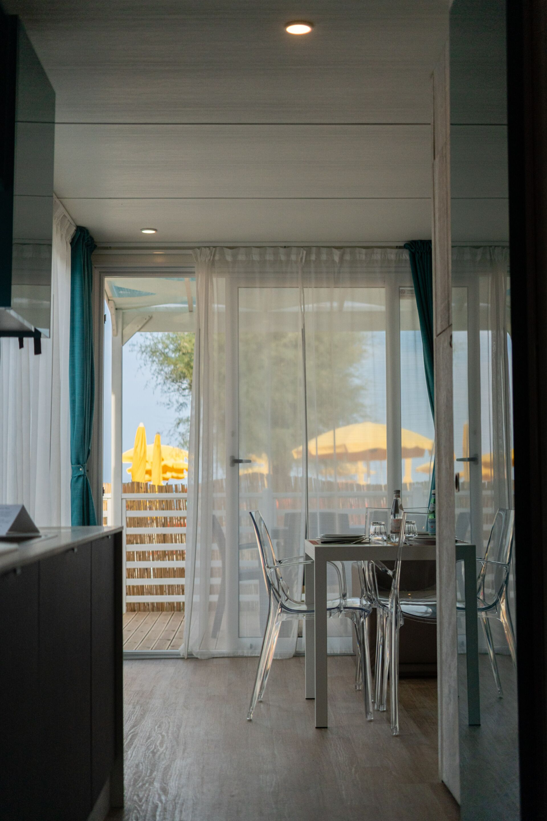 Un espace repas moderne avec une table et des chaises en verre, une vue sur l’espace extérieur grâce à de grandes fenêtres et un plancher en bois.