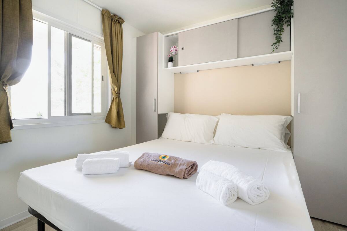 A neatly made bed with white and brown towels, a window with curtains, and a plant on the wall.