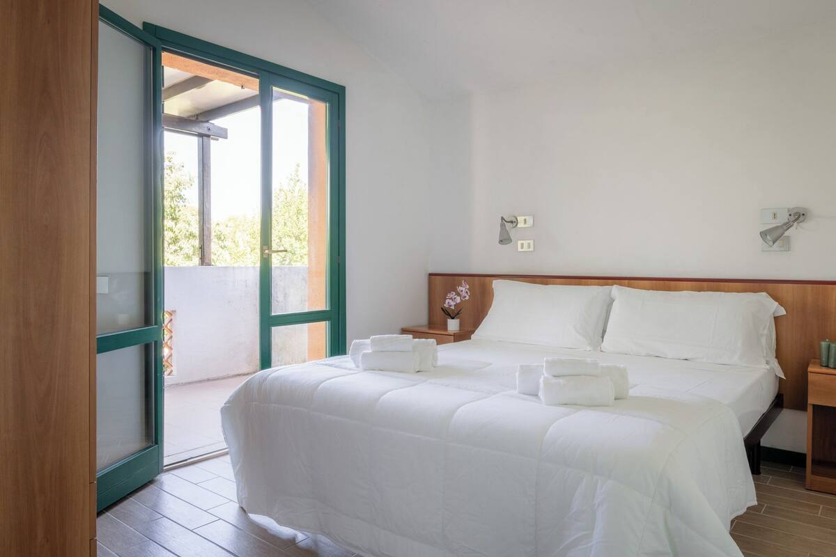 A hotel room with a large bed, a nightstand, a wall-mounted lamp, and a sliding glass door leading to an outdoor area.