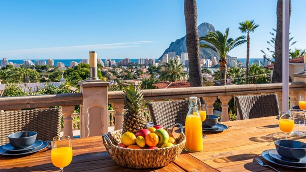 A table set for breakfast with fruit, juice, and a view of a cityscape and mountains. - Villa Suenios - PlusHolidays (Calpe)