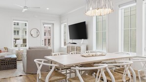 A spacious living room with a large dining table, white furniture, and a television mounted on the wall. - Beach Side Home, Steps from the Sand. Ocean Edge 05 | Bright Side by Kaiser Vacation Rentals. (Orange Beach)