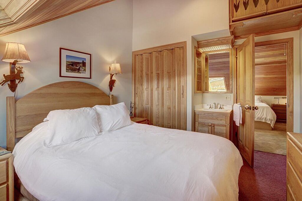A bedroom with a large bed, wooden headboard, two bedside lamps, a framed picture on the wall, a wooden wardrobe, and a bathroom visible through an open door.