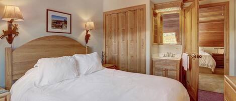 A bedroom with a large bed, wooden headboard, two bedside lamps, a framed picture on the wall, a wooden wardrobe, and a bathroom visible through an open door.