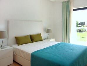 A bedroom with a bed, two pillows, a bedside table with a lamp, and a window with curtains.