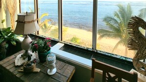 Een kamer met een groot raam dat uitkijkt op een strand, een houten tafel met diverse voorwerpen, en een stoel.