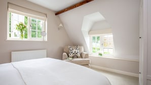 A bedroom with a large bed, a chair with a patterned cushion, and two windows with views of greenery.
