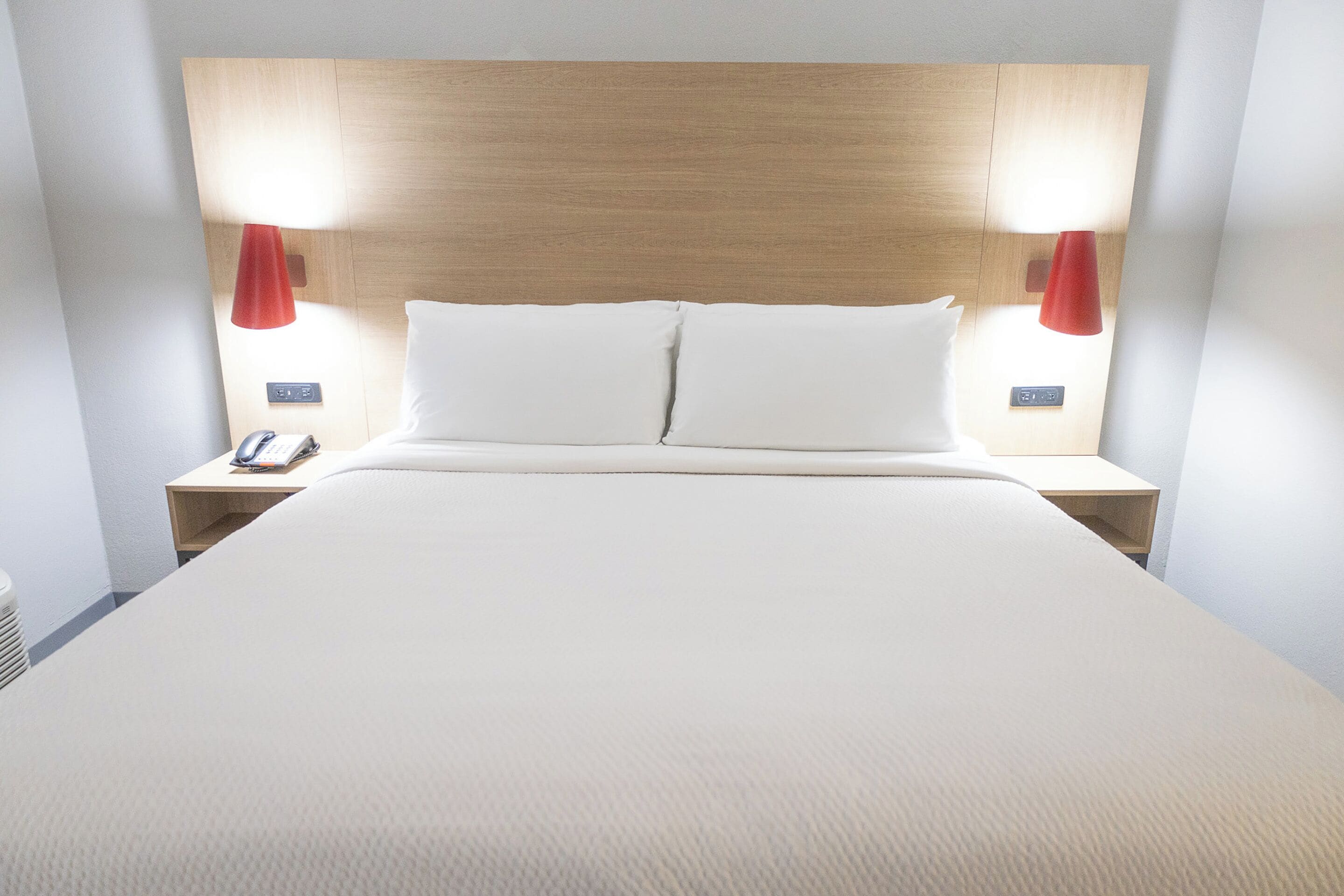 Een net opgemaakt bed met een houten hoofdeinde, twee nachtkastjes met lampen, en een aan de muur bevestigde telefoon.