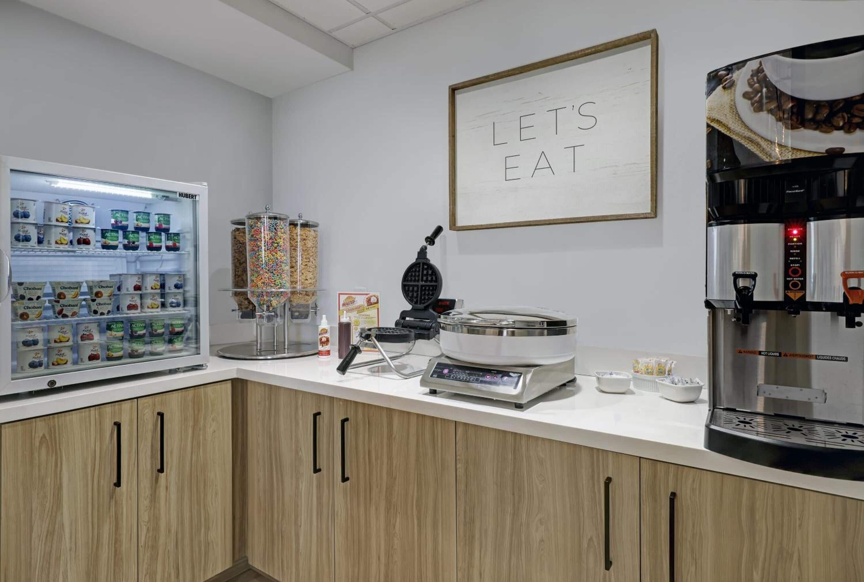 A kitchenette with a refrigerated display case, a waffle maker, a coffee machine, and a 'Let's Eat' sign.