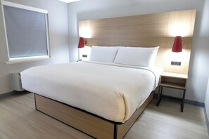 A modern hotel room with a large bed, wooden headboard, two bedside tables with red lamps, and a window with blinds.