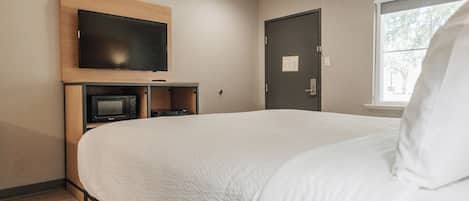 A hotel room with a large bed, a flat-screen TV mounted on a wooden shelf, a microwave, and a door with a window view.