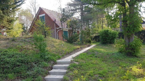A paved pathway leading to a brick house with a sloped roof, surrounded by lush greenery and trees. - House (Condette)