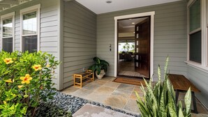 A house exterior with a grey siding, a welcome mat, a small wooden bench, and a potted plant. - Holua Kai #27: Luxury Oceanfront Villa w/Private and Resort Pools & Spas (Kailua Kona)