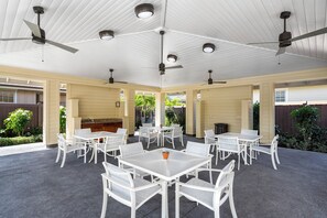 Un patio al aire libre con mesas y sillas blancas, ventiladores de techo y una zona de barbacoa.