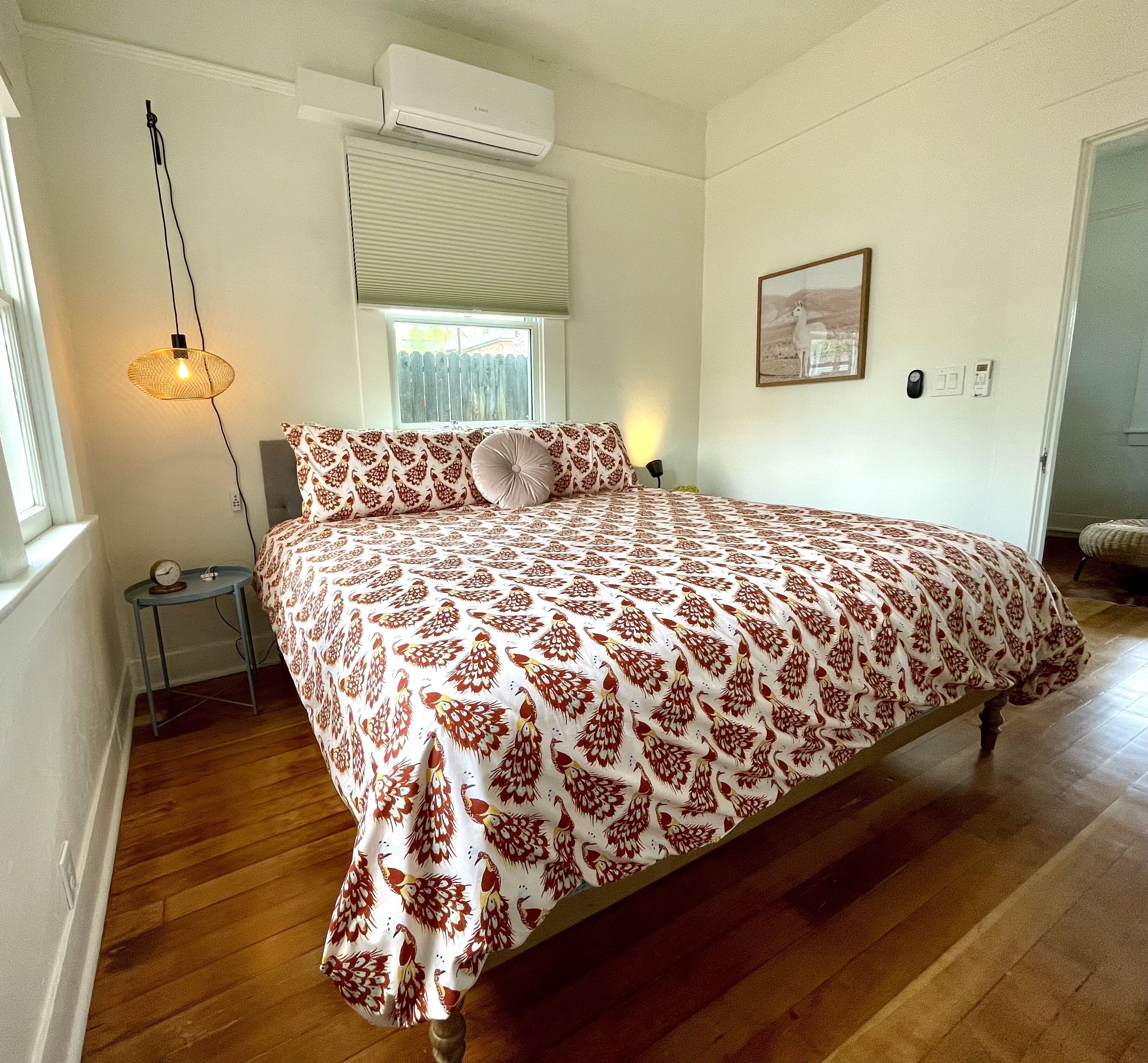A bedroom with a large bed, a bedside table with a lamp, a window with blinds, an air conditioning unit, and a framed picture on the wall.