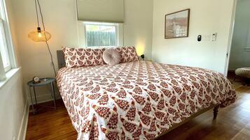 A bedroom with a large bed, a bedside table with a lamp, a window with blinds, an air conditioning unit, and a framed picture on the wall.