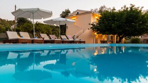 A swimming pool with lounge chairs and umbrellas, a building with a stone facade, and trees in the background. - Villa Sun Sole (Corfu)