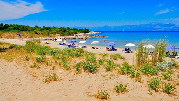 A beach with rows of sunbeds and umbrellas, a clear blue sea, and a distant sailboat. - Villa Sun Sole (Corfu)