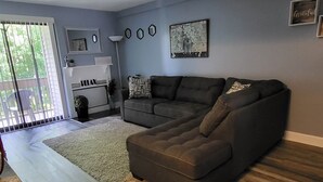 A living room with a sectional sofa, a large window with blinds, a floor lamp, and a wall-mounted artwork. - Spacious 2 Bdrm Condo Walking distance to Weirs Beach!! (Laconia)
