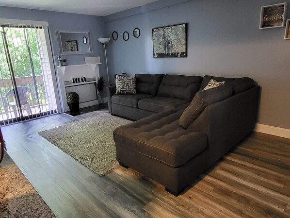 A living room with a sectional sofa, a large window with blinds, a floor lamp, and a wall-mounted artwork.