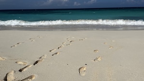 Una playa con huellas que conducen al agua, cielo despejado y olas turquesas.