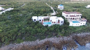 Vista aérea de una propiedad costera con varias construcciones, incluyendo una casa grande con piscina y una edificación más pequeña con techo azul.