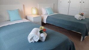 Une chambre d’hôtel avec deux lits, chacun recouvert d’une couverture bleue et agrémenté d’oreillers blancs. Une table de chevet avec une lampe se trouve entre les deux lits, et une serviette blanche ornée de motifs floraux est posée sur chaque lit.
