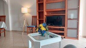 Un salon avec un téléviseur posé sur un meuble en bois, une table basse en verre ornée d’un vase de fleurs, une lampe et un fauteuil.