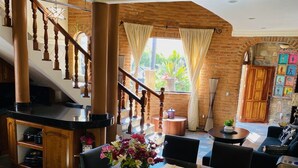 A spacious dining area with a long wooden table, chairs, and a vase of flowers. A staircase with wooden railings leads to an upper level.